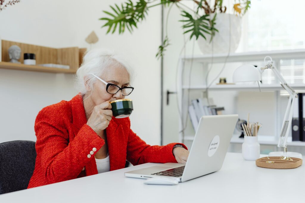 Elderly women wearing a red jacket and drinking coffee, sitting at a desk working on a laptop.