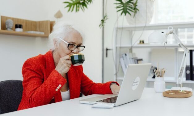 Elderly women wearing a red jacket and drinking coffee, sitting at a desk working on a laptop.