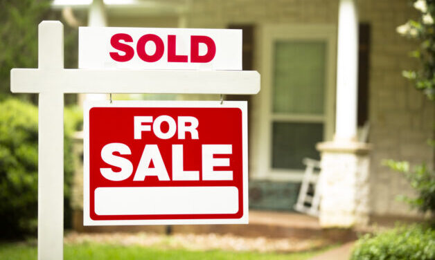 Red and white "Sold, Home for Sale" sign in front of a stone, wood house that is for sale and has recently been sold. Green grass and bushes indicate the spring or summer season. Front porch and windows in background. Real estate signs in residential neighborhood. Moving house, relocation concepts.