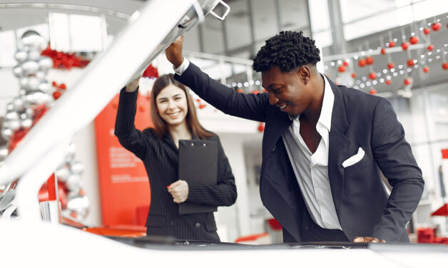 Salesperson opening hood of white car, showing interested customer. There are red and silver balloons in the background.